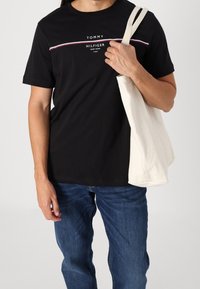 Mand iført en sort Tommy Hilfiger t-shirt og blå jeans, bærer en hvid lærredstotetaske over sin skulder.