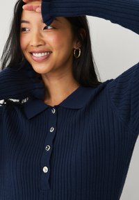 Marineblaues geripptes Poloshirt mit Kragen, ausgestattet mit silbernen Knöpfen auf der Vorderseite und langen, strukturierten Ärmeln.