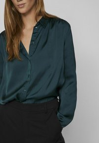 Blouse en satin vert d'eau avec un devant boutonné, un col et des manches longues, associée à un pantalon noir. Texture lisse et coupe décontractée.