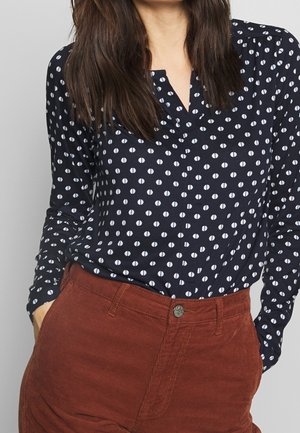 Femme portant un haut à manches longues bleu marine avec un motif en demi-cercle blanc, rentré dans un pantalon en velours côtelé taille haute de couleur rouille, les mains dans les poches.