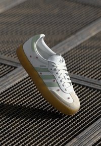 Baltas sporta apavi ar zaļiem akcentu svītrainēm, ziedu detaļām, teksturētu gumijas zoli un gludu augšpusi. Dizains ietver klasiskās trīs svītru zīmola iezīmes.