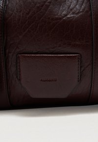 Saco de couro bordeaux escuro com uma superfície texturada e um bolso cosido com uma marca em relevo. Forma arredondada com um acabamento polido.