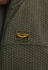 Oliwkowa kurtka z teksturowanej tkaniny, z wyraźną żółto-czarną metką, na której widnieje napis "PME LEGEND, AMERICAN CLASSIC." Widziany zamek.
