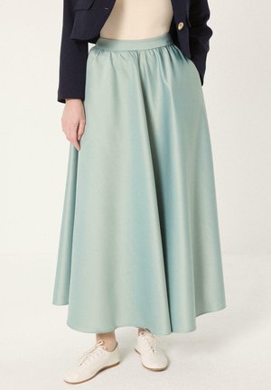 Femme portant une longue jupe en satin bleu clair, des baskets blanches, un haut crème et un blazer bleu marine avec un bouton doré, debout sur un sol blanc.