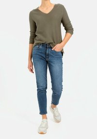 Długi rękaw sweter w kolorze oliwkowym z dekoltem w kształcie V połączony z niebieskimi dżinsami. Obuwie to szare i białe sneakersy z gumową podeszwą.