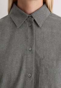 Γκρι φλάνελ πουκάμισο με κλασικό γιακά, εμπρόσθια κουμπότρυπα με γκρι κουμπιά και τσέπη στο στήθος, που επιδεικνύει έναν μαλακό, υφασμάτινο υφή.