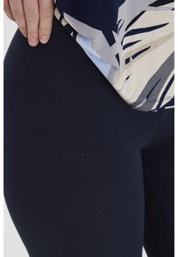 Jūriniai legginsai iš elastingos medžiagos, derinami su šviesiu raštuotu marškinėliu, kuriame vyrauja mėlynos ir neutralios spalvos, pabrėžiančios dinamiškus dizainus ir formas.