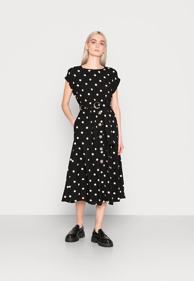 Schwarzes Kleid mit Polka-Punkten und tailliertem Schnitt, mit kurzen Ärmeln, einem Gürtel und Knöpfen an der Vorderseite, kombiniert mit klobigen schwarzen Schuhen.