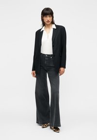 Blazer noir oversize, haut blanc à col, jeans évasés gris foncé et talons imprimé léopard. Textures lisses et éléments de design sur mesure.