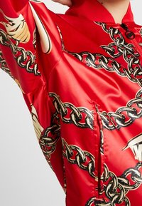 Röd bomberjacka med en glansig yta, med ett kedjemönster i svart och guld. Inkluderar dragkedjest ängning och ribbad krage.