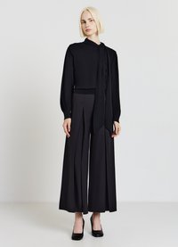 Donna con bob biondo che indossa una blusa nera a maniche lunghe con cravatta e pantaloni neri a palazzo plissettati con tacchi neri.