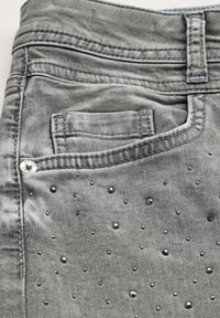 Grijze denim jeans met een gestructureerd oppervlak, voorzien van kleine decoratieve zwarte studs en een voorzak. Gevoerde details en een metalen knoop zijn zichtbaar.
