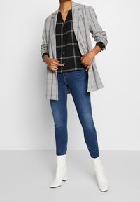 Grå rutig kavaj över en svart rutig skjorta, matchad med blå smala jeans och vita klackboots. Släta texturer och en strukturerad design.