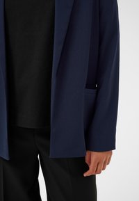 Tumši zils bleiseris ar gludu tekstūru, ar vienu pogu aizdari un sānu kabatām, kārtots pār melnu krekla, kopā ar melnām biksēm.