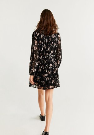 Frau geht weg, trägt ein schwarzes, florales Langarm-Minikleid mit plissiertem Rock und schwarzen flachen Schuhen auf weißem Hintergrund.