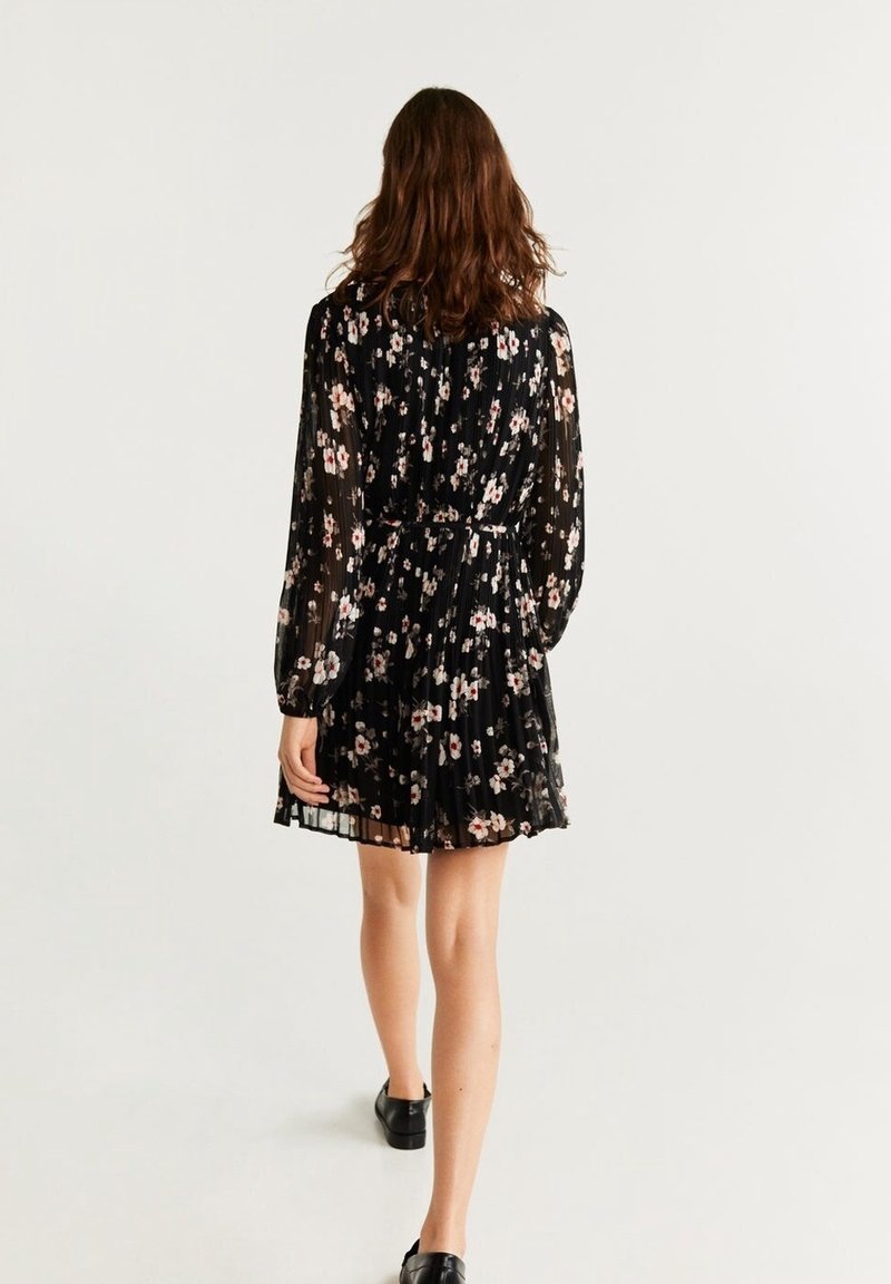 Femme qui s'éloigne portant une mini-robe noire à fleurs à manches longues avec une jupe plissée et des chaussures plates noires sur fond blanc.