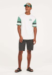 T-shirt blanc avec des manches rayées vertes et noires, arborant le mot "PRO TEST" en vert. Accompagné d'un short gris foncé et de sandales noires.