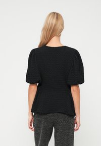 Blusa preta texturizada com mangas bufantes curtas, decote redondo e formato peplum, usada sobre calças escuras com padrão.
