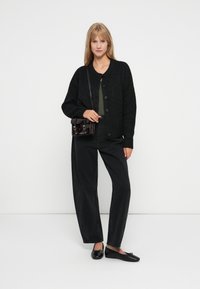Svart strikket cardigan med knapper, grønn topp, svarte vide bukser og svarte leiligheter. Liten mørk håndveske med doble låser. Ren bakgrunn.