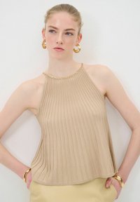 Beige geribbelde gebreide top met een halternek ontwerp en een gouden kettingaccent. De stof lijkt zacht met een vloeiende vorm en zijsplitten.