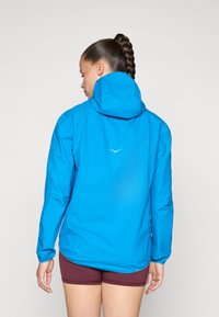 Woman with braided hair wearing a bright blue hooded jacket and maroon shorts, standing facing away against a plain background.