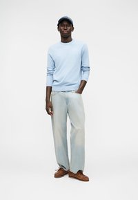 Pull bleu clair, jean bleu clair et chaussures en cuir marron. La personne se tient avec confiance, portant une casquette bleue assortie. Look simple et décontracté.