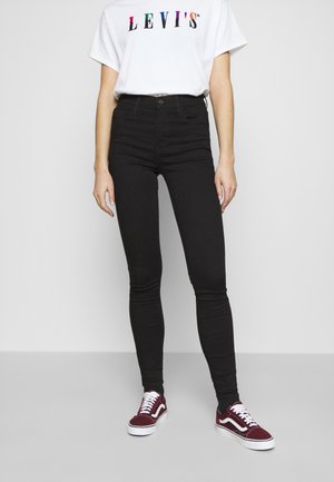 Jean skinny noir taille haute, associée à un t-shirt blanc arborant un logo Levi’s multicolore et des baskets bordeaux.