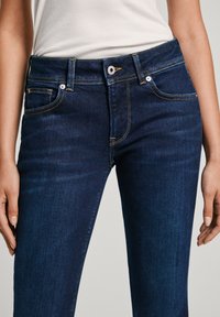 Person som bär mörkblå denimjeans och en vit ribbad ärmlös topp, står med armarna avslappnat vid sidorna.