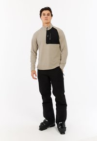 Lichtgrijze pullover met zwarte borstzak en een halve ritssluiting. In combinatie met zwarte skibroek en zwarte skibotten, wat een sportieve uitstraling laat zien.