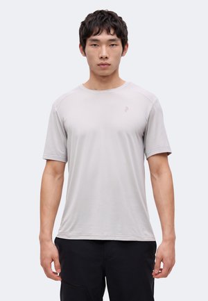Jeune homme de face portant un t-shirt de sport gris clair à manches courtes et un pantalon noir sur un fond uni.
