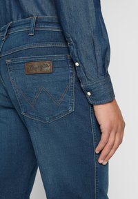 Jean en denim bleu foncé avec un design de poche arrière distinctif, comportant un patch en cuir Wrangler et des coutures orange contrastantes.