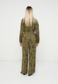 Donna con lunghi capelli biondi lisci girata di spalle, che indossa una tuta floreale verde oliva e scarpe scure su uno sfondo bianco.