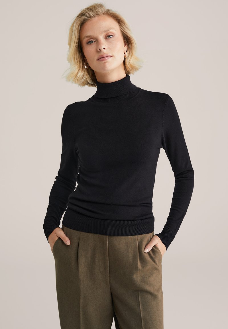 Juodas golfas prigludusiu siluetu su ilgomis rankovėmis, raštuotais kraštais ir minkšta tekstūra, derinamas su aukštu liemeniu rudomis kelnėmis.