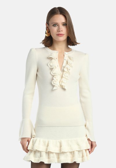 Kermanvärinen, ribbikuvioitu neulemekko, jossa on röyhelökaulus, nappikoristeet ja liehuvat hihat; kerroksittainen röyhelöhelma lisää tekstuuria.