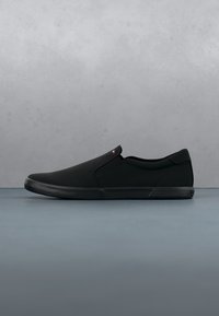 Basket noire basse profil sans lacets avec semelle en caoutchouc et petit logo rouge et blanc près du panneau latéral élastique, sur une surface grise avec un fond gris uni.