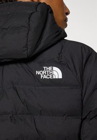 Svart puffjacka med en texturerad, vadderad design. Har en framträdande vit logotyp på övre ryggen och en huva. Mjuk materialfinish.