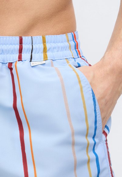 Κοντινό πλάνο ενός ατόμου που φοράει ανοιχτό μπλε σορτς με κάθετες πολύχρωμες ρίγες και ελαστική μέση, με το χέρι να ακουμπά στην τσέπη.