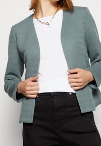 Blazer vert menthe texturé avec un motif en chevrons et un design à ouverture avant, porté sur un t-shirt blanc et un pantalon noir, les mains saisissant les revers.