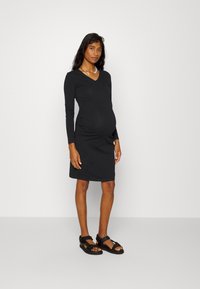 Vestido de maternidade preto de manga longa com decote em V e laterais pregueadas, combinado com sandálias pretas com correias ajustáveis e sola texturizada.