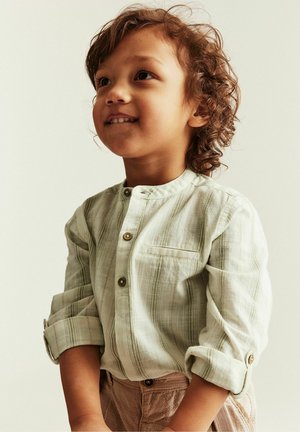 Young child with curly hair wearing a light striped buttoned shirt and beige pants, smiling and looking slightly upward.