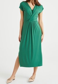 Zelené midi šaty s krátkými balónovými rukávy, výstřihem do V a zkrouceným detailem vpředu. Hladký materiál, postranní rozparek, v kombinaci s světlými balerínami.
