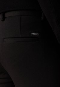 Zwarte op maat gemaakte broek van textuurstof, met een vlakke voorkant, zijzakken en een klein merklabel op de achterzak.
