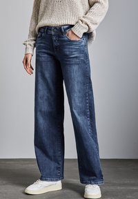 Jeans de mezclilla azul de pierna ancha con un acabado desgastado, que cuentan con bolsillos frontales y un cierre de botón, combinados con un suéter ligero de textura y zapatillas blancas.