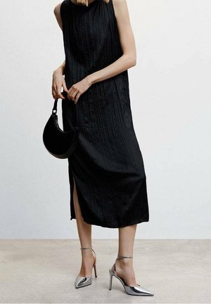 Femme portant une robe midi noire sans manches avec une fente sur le côté, tenant un sac à main noir et chaussée de chaussures à talons aiguilles argentées.