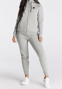 Grijze zip-up hoodie met trekkoorden, voorzakken en een geborduurd logo, gecombineerd met bijpassende grijze joggers en witte sneakers.