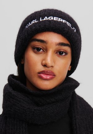 Sort strikket beanie med "KARL LAGERFELD" broderet i hvid, kombineret med et matchende struktureret tørklæde. Glat hudtone, neutral baggrund.