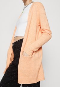 Osoba v broskvově zbarveném otevřeném cardigan s kapsami, bílém cropped topu a černých džínech s vysokým pasem a hvězdičkovým vzorem.