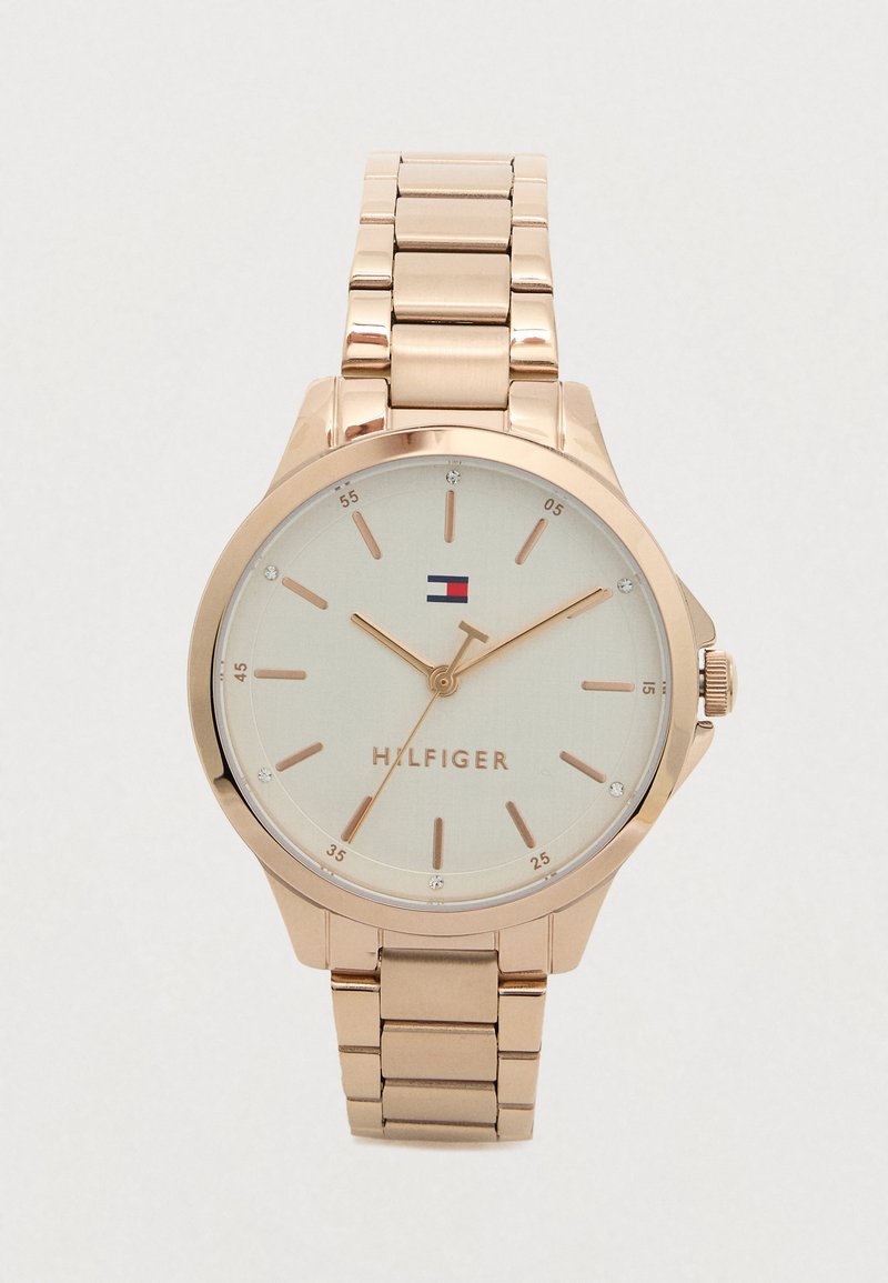 Námořnické hodinky z růžového zlata s propojeným páskem, béžovým ciferníkem, minimalistickými hodinovými značkami a logem Tommy Hilfiger na pozici 12 hodin.