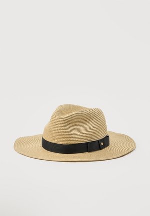 Beigenvärinen kudottu leveälierinen fedora-hattu, jossa musta nauha ja pieni metallinen logo yksivärisellä vaalealla taustalla.