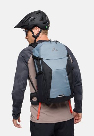 Man met een zwarte fietshelm, zonnebril en een shirt met lange mouwen, met een grote blauw-zwarte rugzak voor wandelen tegen een effen achtergrond.